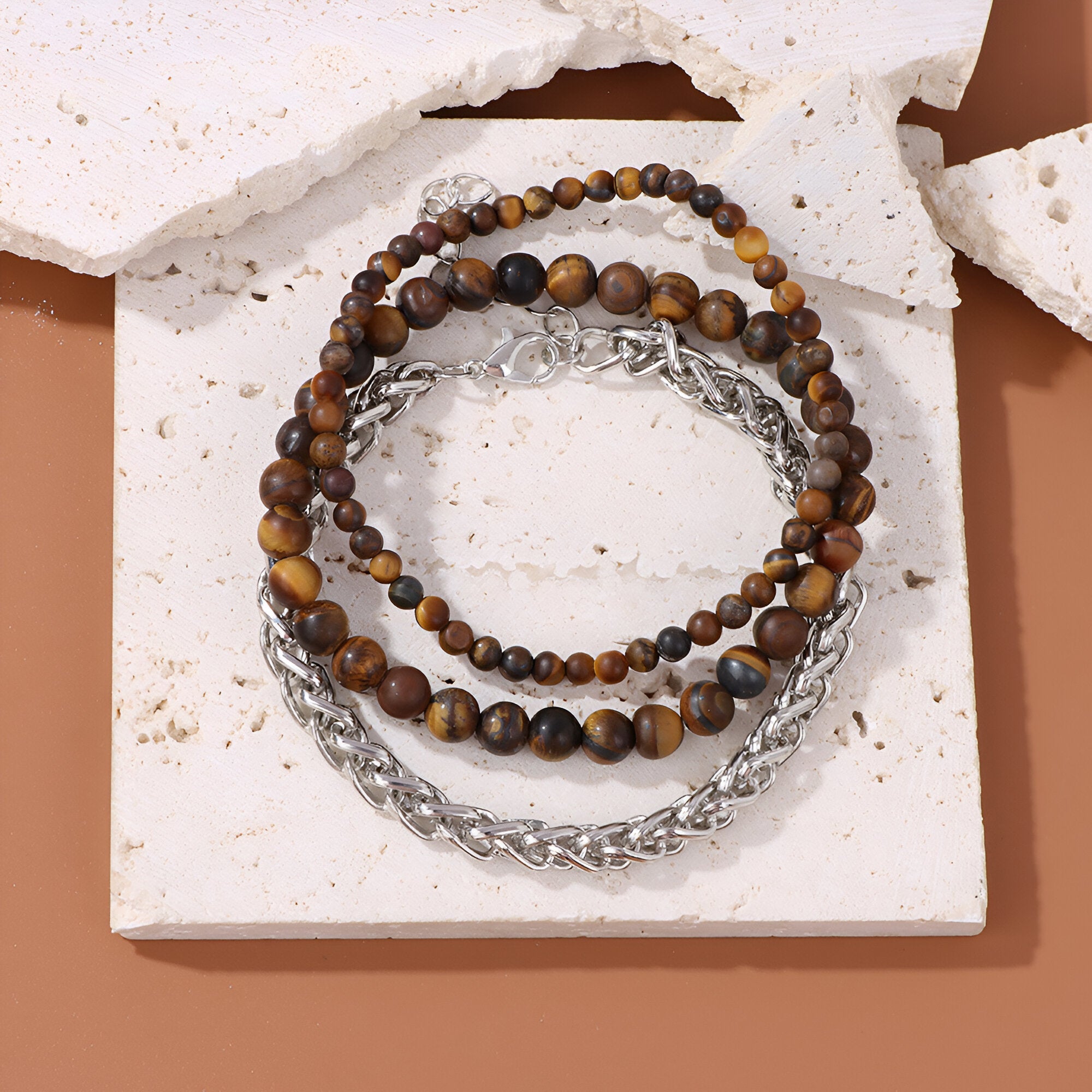 Trio de bracelets pour homme avec perles en œil de tigre et chaînes en acier argenté sur fond beige.