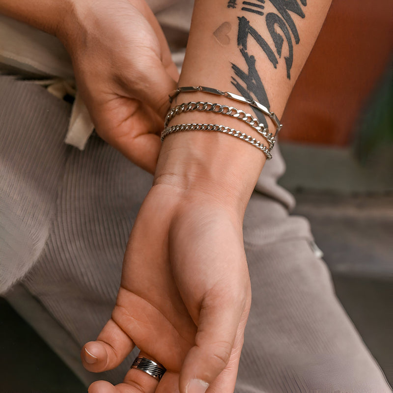 Trio de bracelets maille en titane argent porté sur poignet masculin avec tatouage noir.