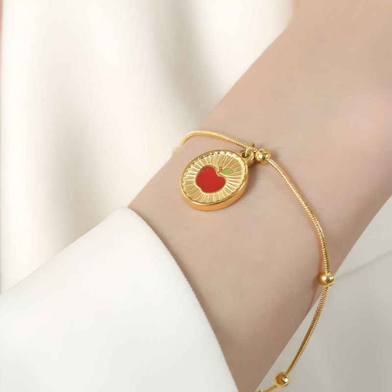 Bracelet fin en titane plaqué or 18 carats avec médaille dorée gravée d'une pomme rouge au centre.