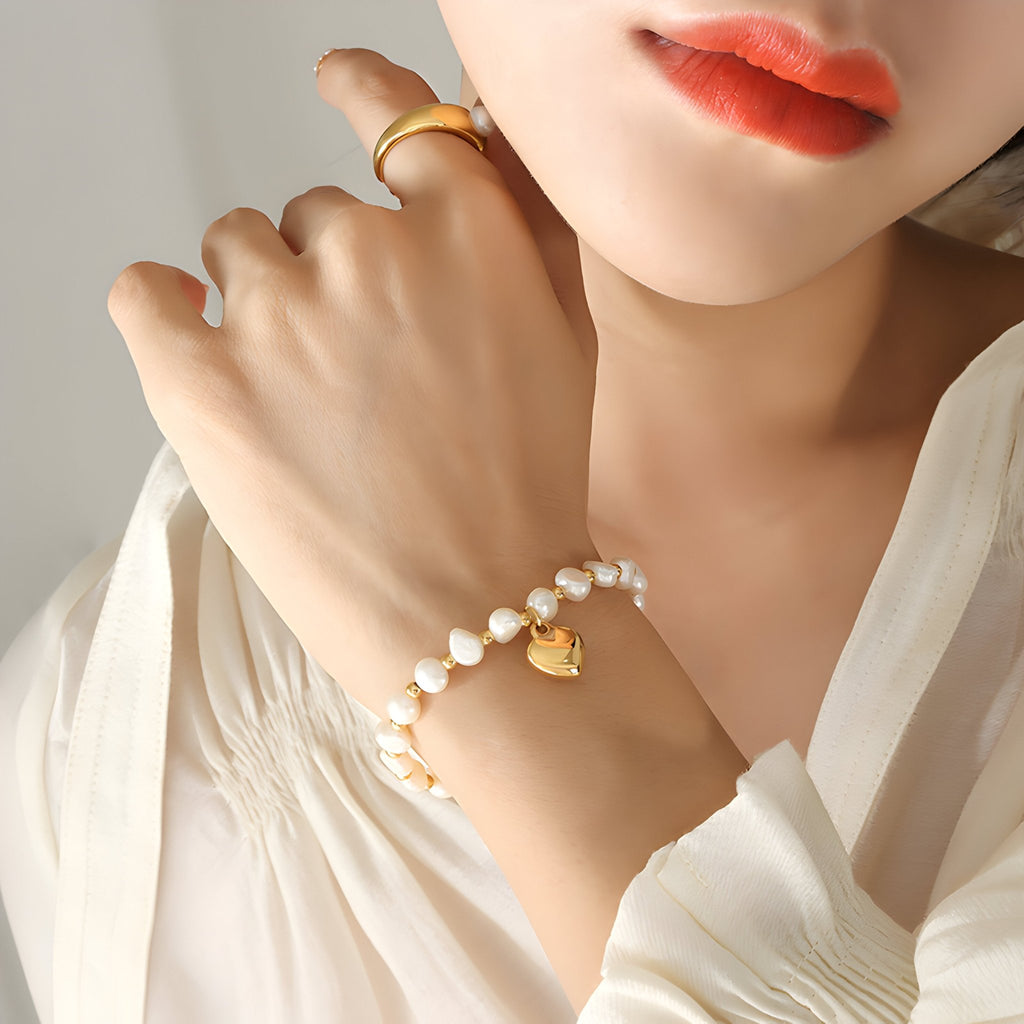 Bracelet en titane plaqué or avec perles blanches et pendentif cœur doré, porté au poignet féminin.
