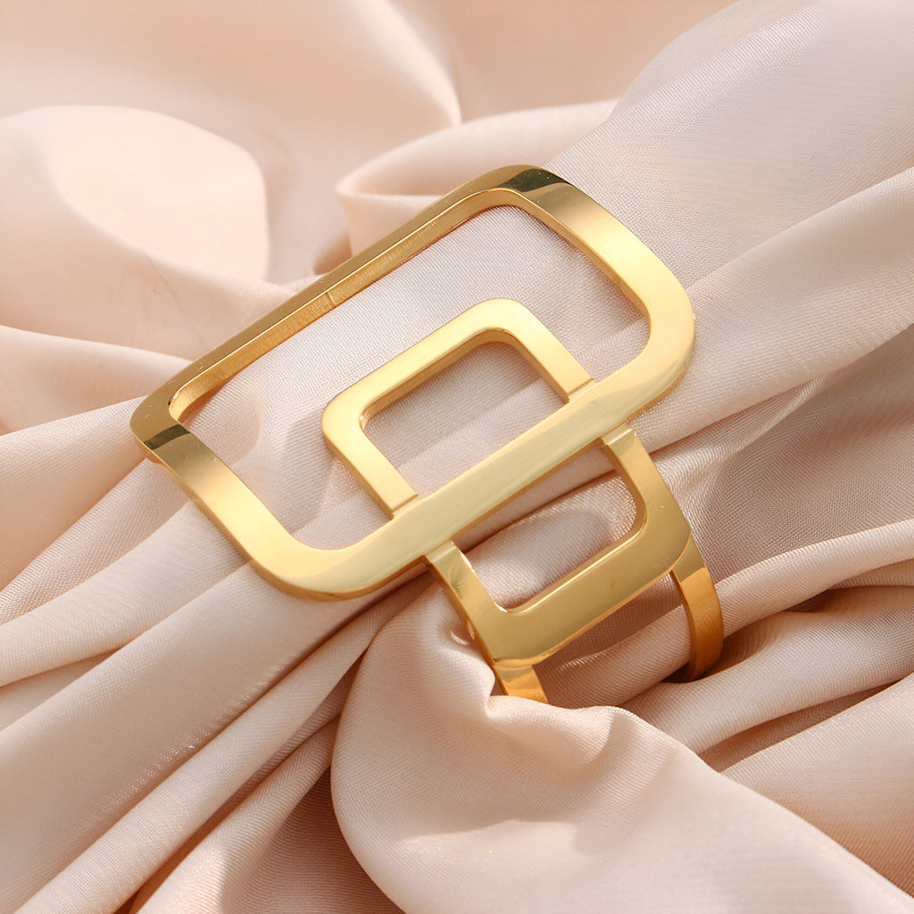 Bracelet manchette doré avec design géométrique carré et finition brillante sur tissu satin crème.
