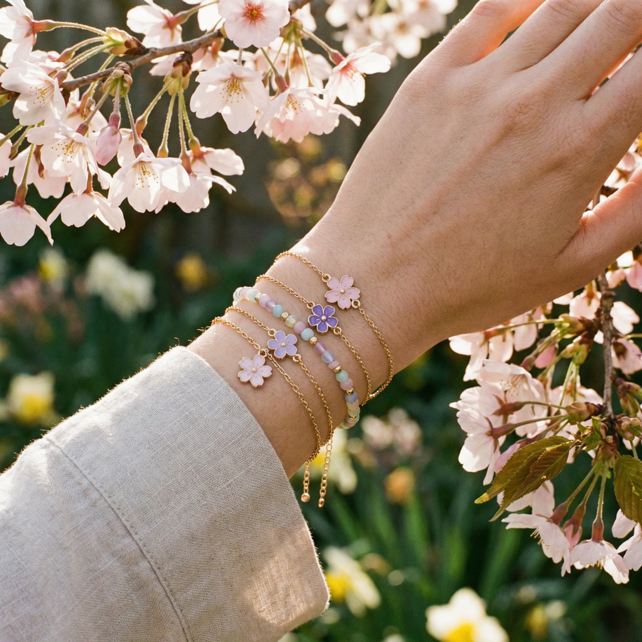 Poignet de femme portant plusieurs bracelets printaniers avec fleurs en émail et perles pastel, cerisiers en fleurs en arrière-plan
