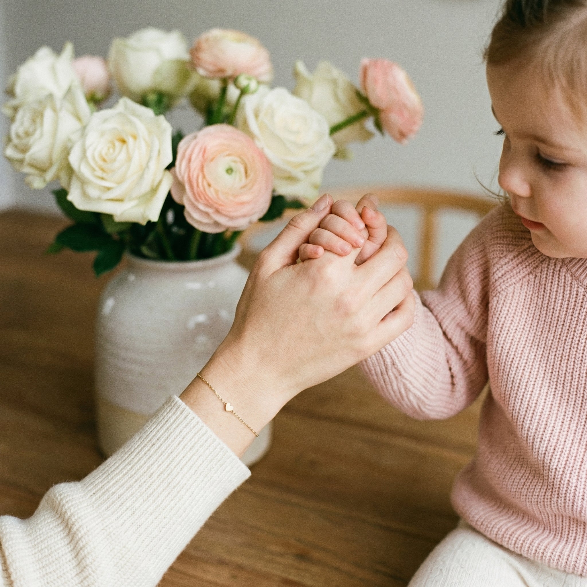 Maman et enfant main dans la main, bracelet doré cœur au poignet, bouquet de roses blanches et roses en arrière-plan