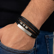 Bracelet en cuir noir multi-rangs avec détails tressés et bijou argenté mat sur poignet.