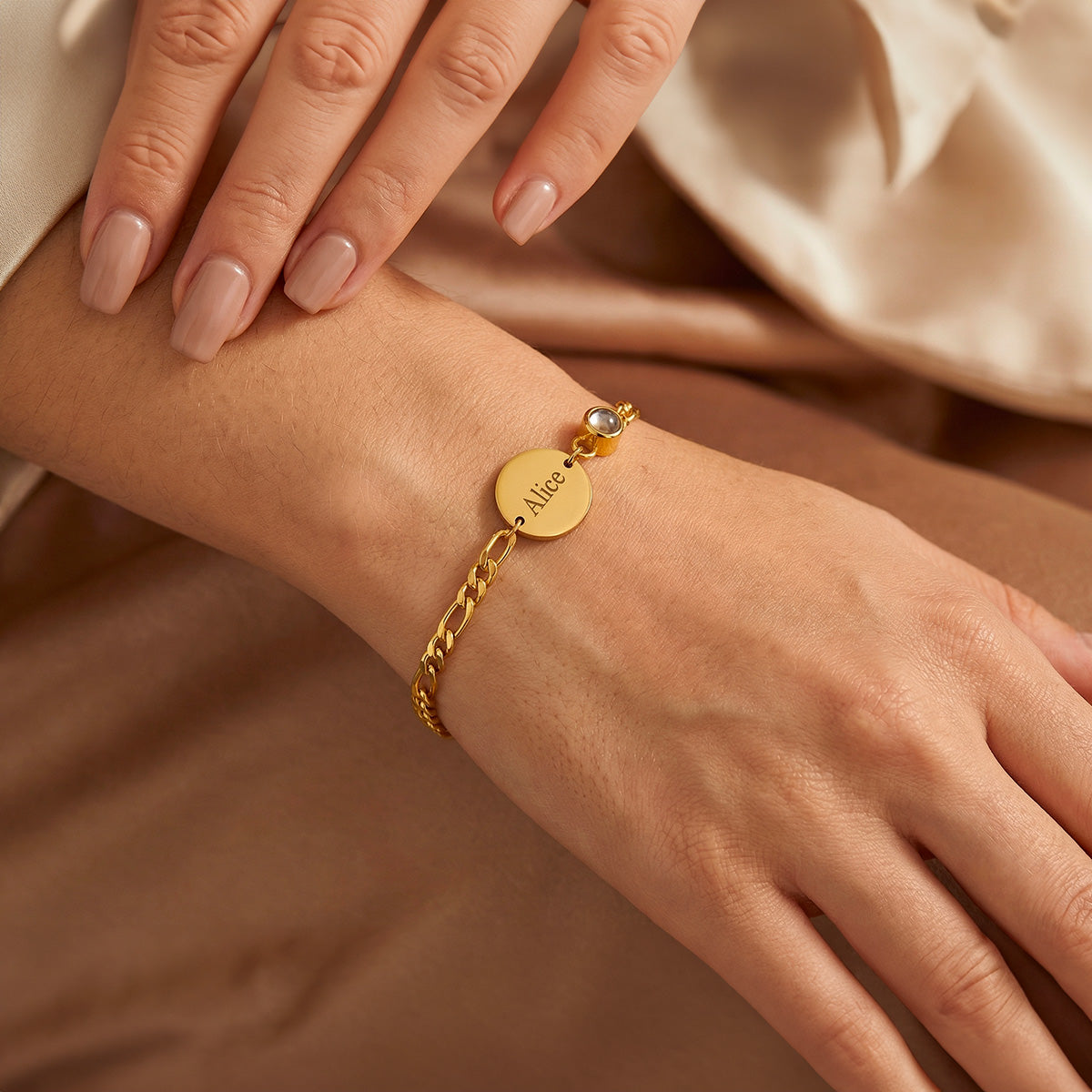 Bracelet en chaîne Figaro doré avec médaille ronde gravée et petit pendentif sur poignet féminin.