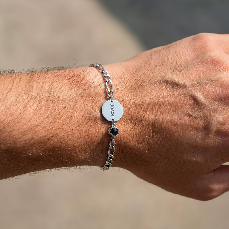 Bracelet en argent avec maille figaro, médaille ronde gravée et petit pendentif noir sur poignet masculin clair.