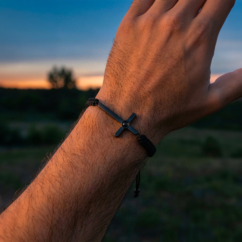 Bracelet noir en corde coulissante avec pendentif croix argentée gravée porté au poignet.