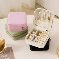 Boîte à bijoux nomade rose avec compartiments intérieurs rembourrés et crochets pour pendentifs, placée sur une boîte verte.