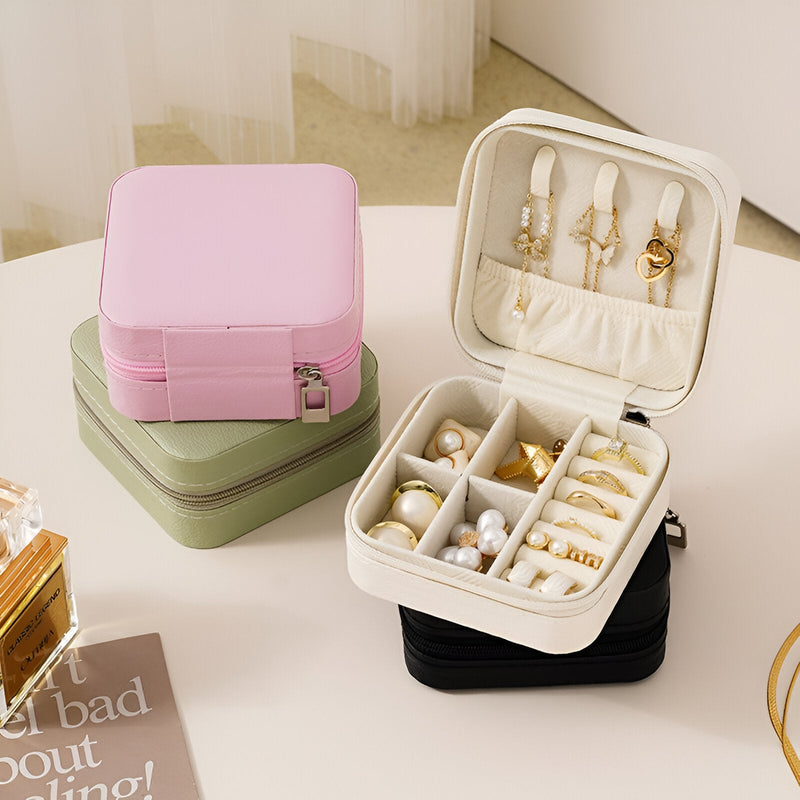 Boîte à bijoux nomade rose avec compartiments intérieurs rembourrés et crochets pour pendentifs, placée sur une boîte verte.