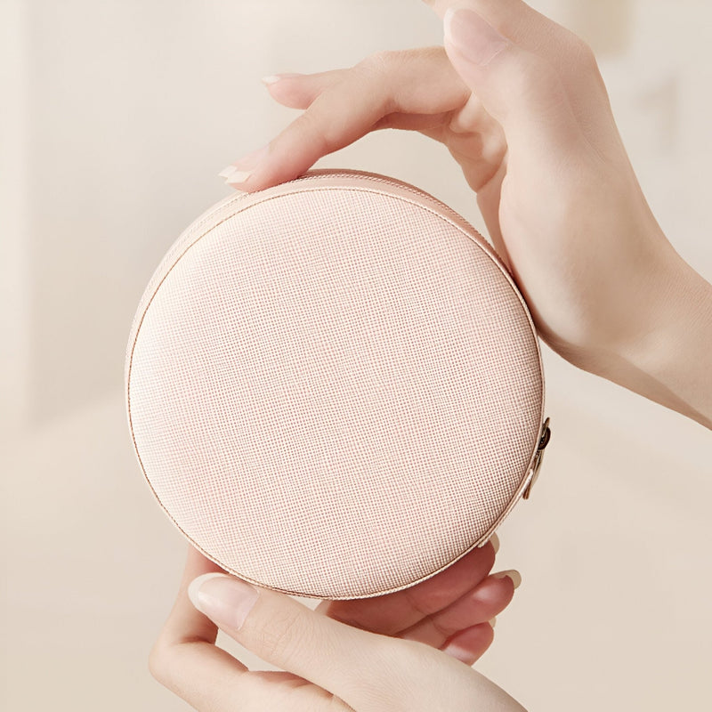 Boîte à bijoux nomade ronde en cuir rose pâle avec fermeture éclair dorée.