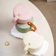 Boîte à bijoux nomade ronde rose avec fermeture bouton doré et anse en cuir rose pâle.