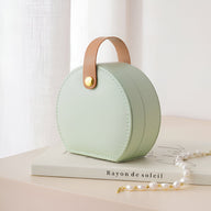 Boîte à bijoux nomade arrondie verte avec anse en cuir camel et bouton doré.