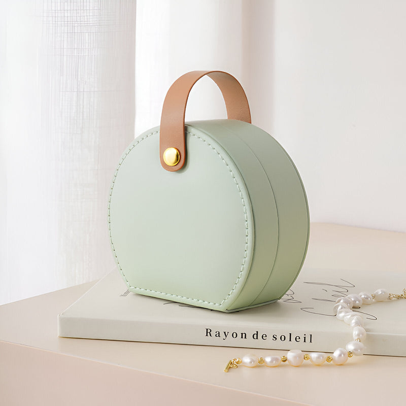 Boîte à bijoux nomade arrondie verte avec anse en cuir camel et bouton doré.