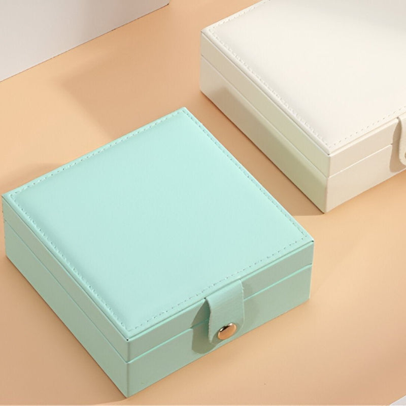 Boîte à bijoux nomade carrée en cuir bleu clair avec bouton-pression doré et boîte blanche en arrière-plan.