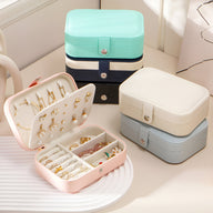 Boîte à bijoux nomade rose ouverte avec compartiments, bagues et boucles d'oreilles, autres boîtes empilées blanc, bleu.