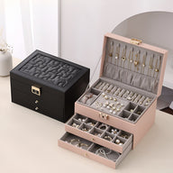 Boîte à bijoux nomade rose à multiples tiroirs ouverts avec compartiments pour bagues et colliers, boite noire fermée à côté.