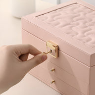 Boîte à bijoux nomade rose à grands tiroirs avec fermeture dorée et dessus matelassé élégant.