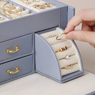 Boîte à bijoux bleue avec tiroirs et rangement incliné pour bagues, mains insérant une bague dorée.