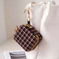 Boîte à bijoux nomade mini à carreaux marron, fermeture éclair jaune, mousqueton doré attaché à un sac blanc.