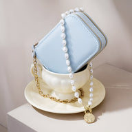 Boîte à bijoux nomade bleue en forme de sac à main, chaîne dorée et collier de perles blanches avec médaillon placé dans une .