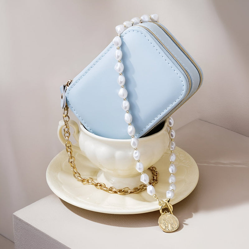 Boîte à bijoux nomade bleue en forme de sac à main, chaîne dorée et collier de perles blanches avec médaillon placé dans une .