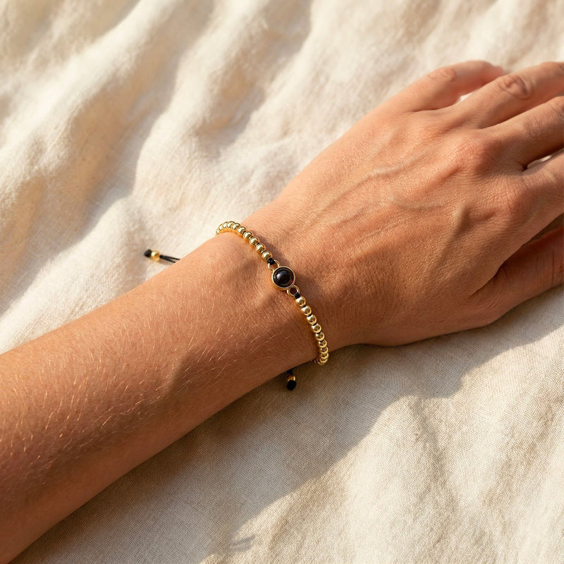 Bracelet à perles métalliques dorées avec perle centrale noire sur cordon noir ajustable porté au poignet.