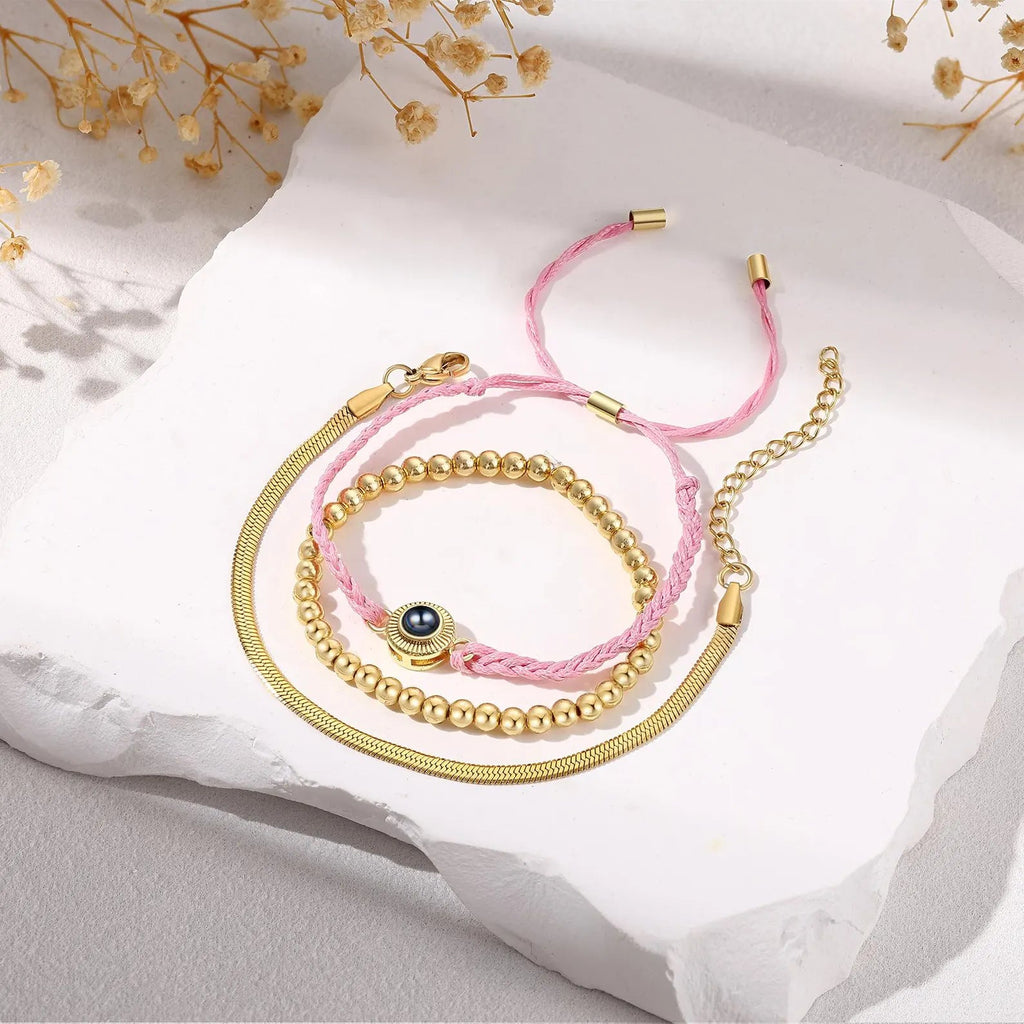 Bracelet trio en acier inoxydable avec perles dorées, cordon tressé rose et pendentif œil bleu.