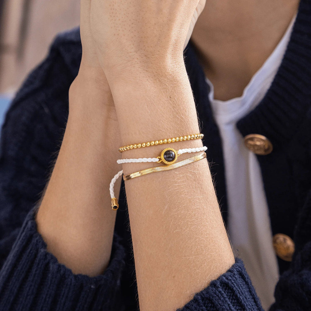 Bracelet à cordon tressé beige avec trio de perles dorées et médaille centrale en acier inoxydable noir brillant.