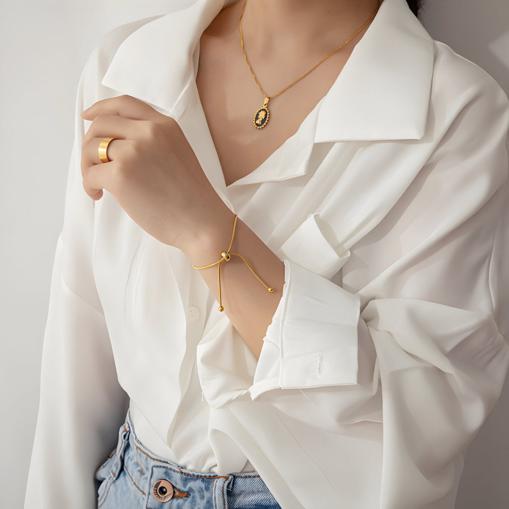 Bracelet ajustable en titane doré avec pendentif ovale et chaîne fine sur poignet féminin vêtu d'une blouse blanche.