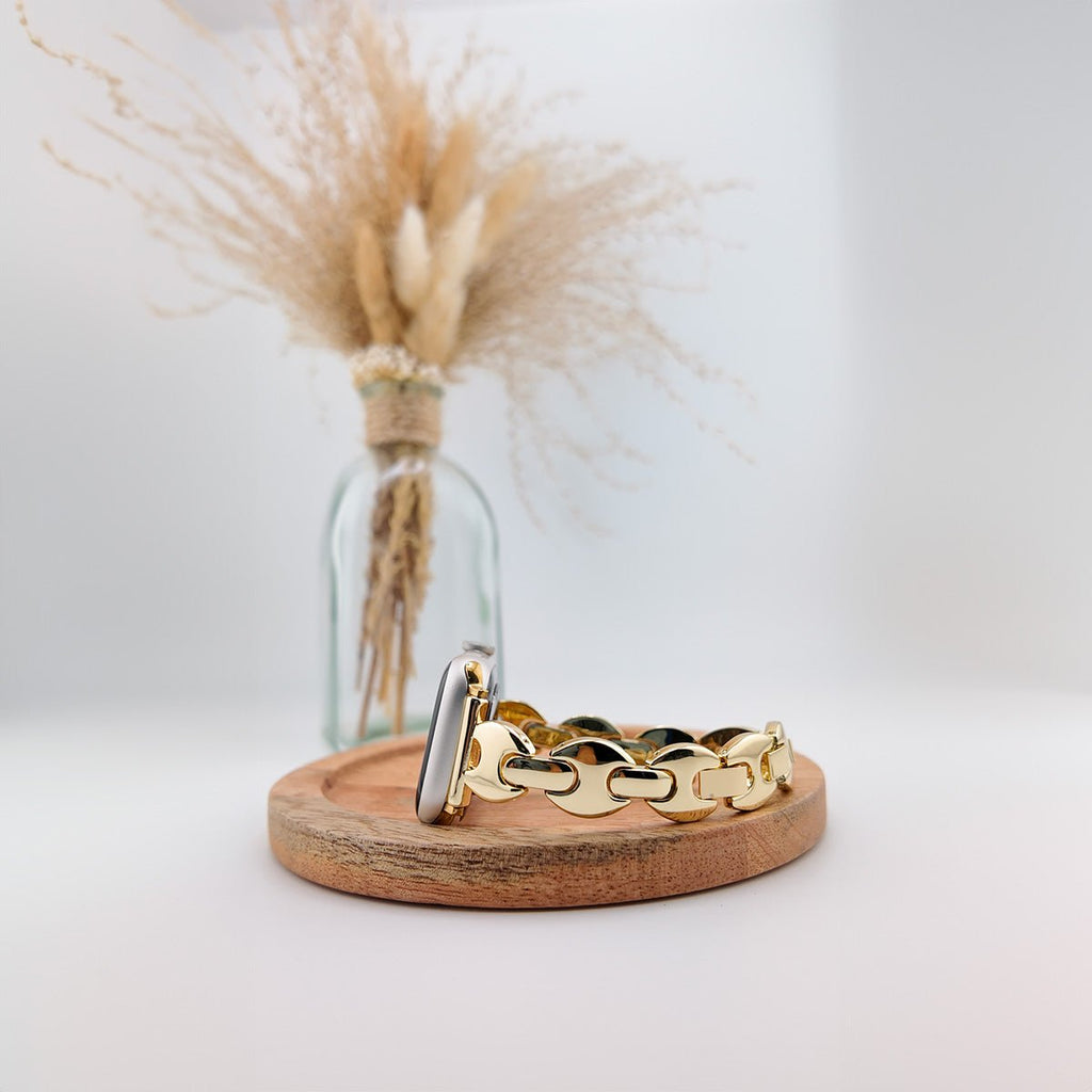 Bracelet Apple Watch à mailles larges dorées avec fermoir argenté posé sur un socle en bois clair.