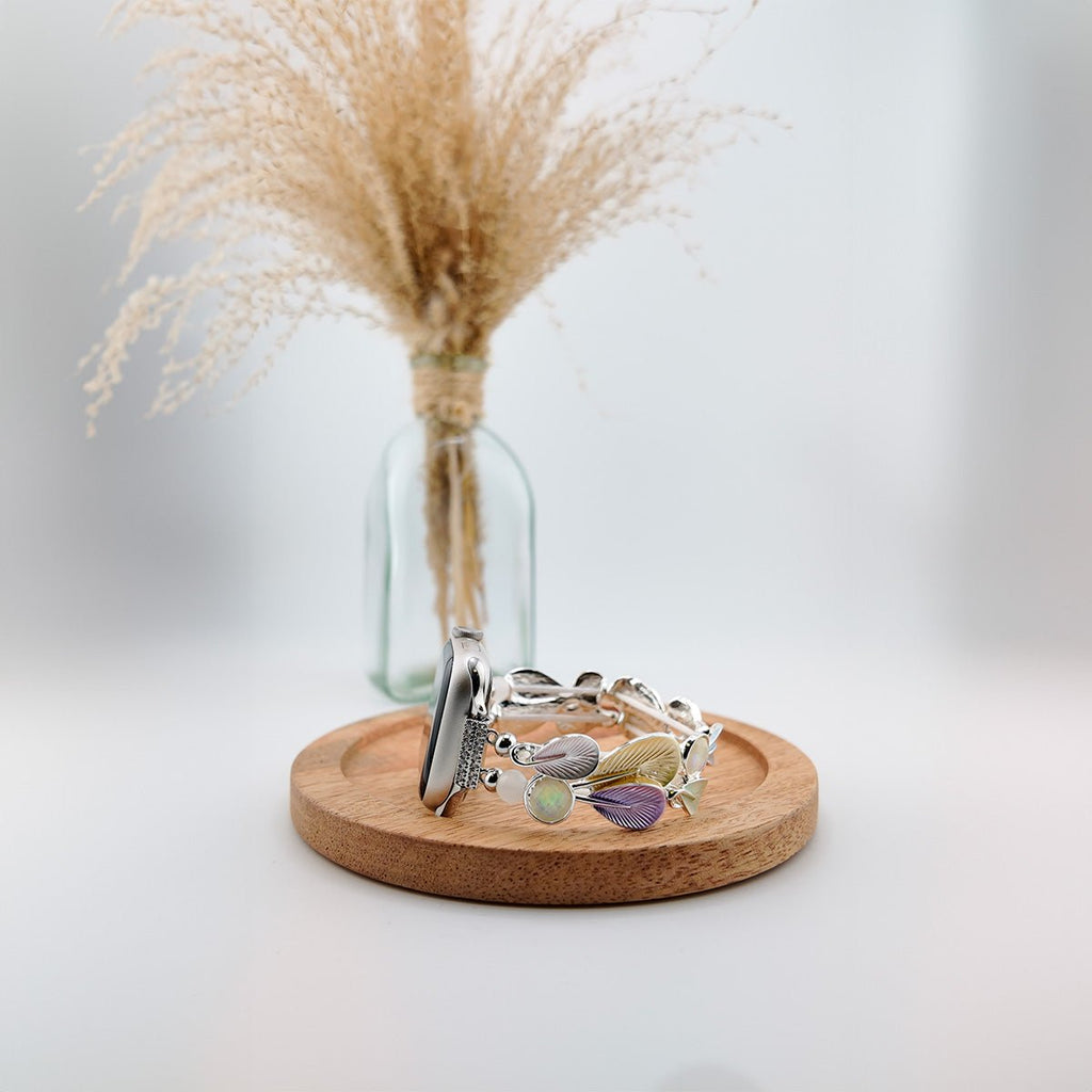 Bracelet Apple Watch femme en métal argenté avec breloques en forme de plumes colorées, posé sur un socle en bois rond.