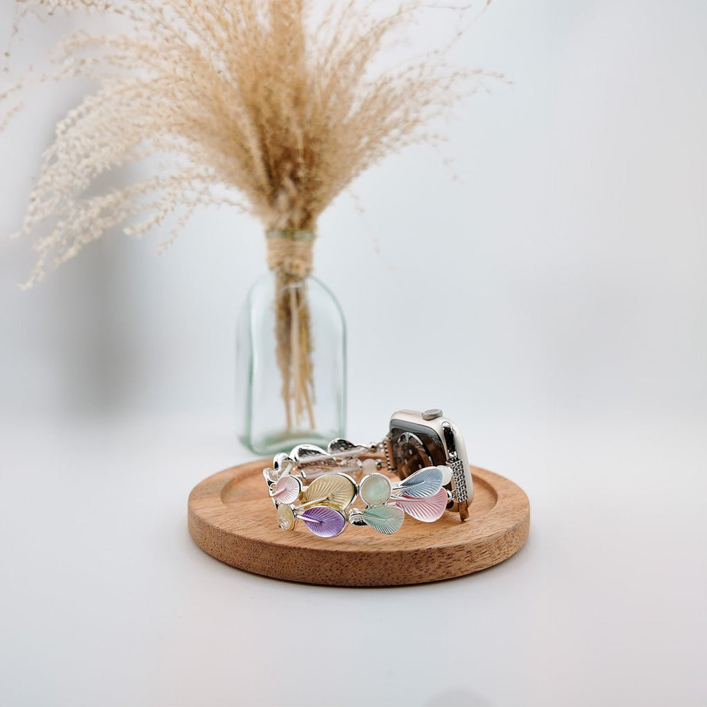 Bracelet Apple Watch pour femme avec breloques plume pastel multicolore sur socle bois rond.
