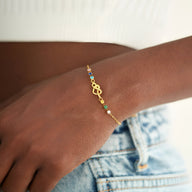 Bracelet fin en or avec pendentif cœur emmêlé et perles colorées sur poignet féminin.