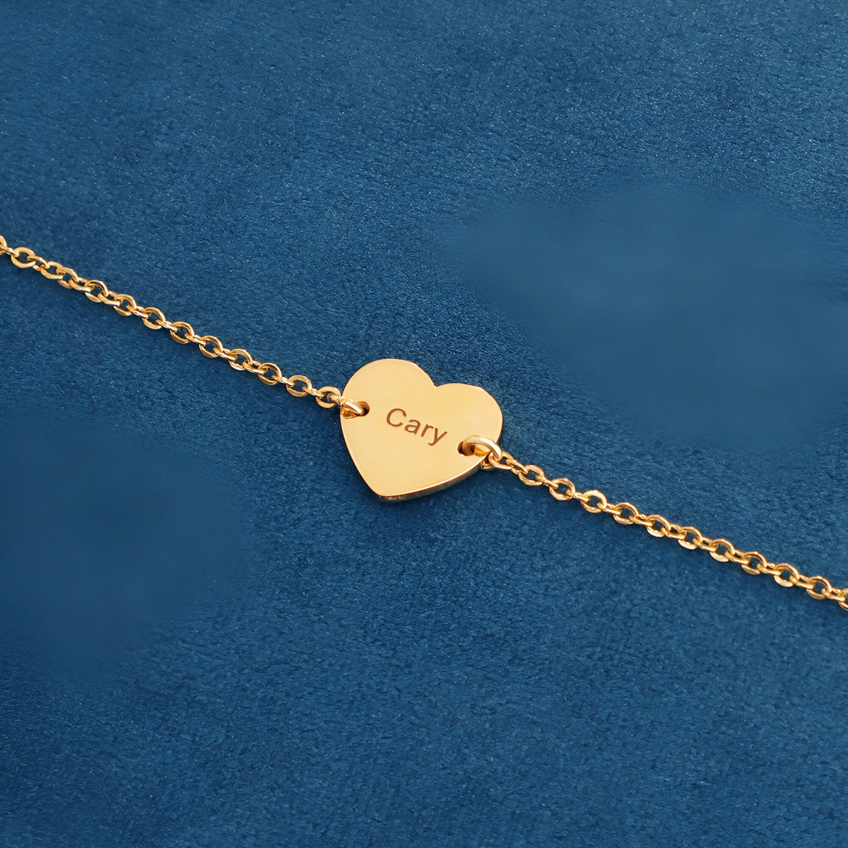 Bracelet fin en acier inoxydable doré avec pendentif cœur gravé « Cary » sur fond bleu.