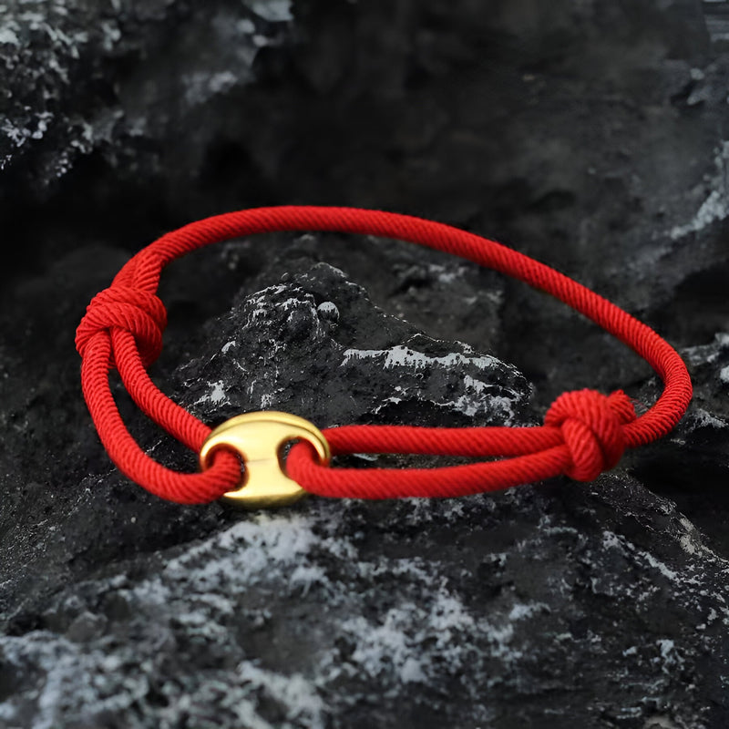 Bracelet cordon rouge avec fermoir métallique doré minimaliste sur fond rocheux noir.