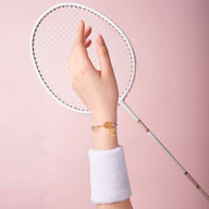 Bracelet cordon or avec pendentif badminton en acier inoxydable porté au poignet femme sur fond rose.
