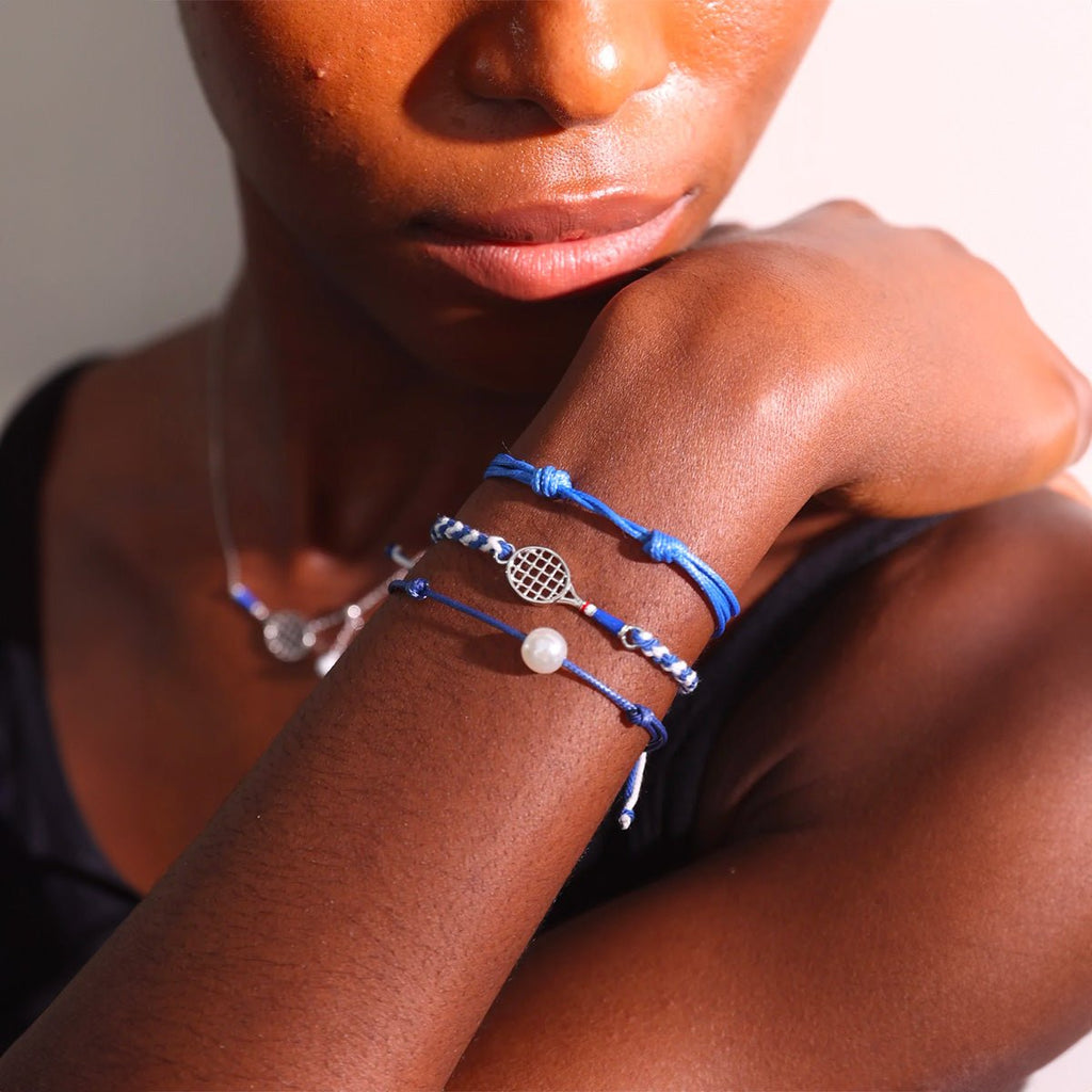 Bracelet cordon bleu avec pendentif forme balle de tennis en acier inoxydable sur poignet peau foncée.