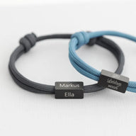 Bracelet cordon noir avec plaque acier inoxydable gravée "Markus Ella" et bracelet cordon bleu clair avec plaque gravée.