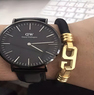 Bracelet cordon noir avec pendentif doré en forme de chiffre 9 porté au poignet.