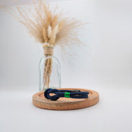 Bracelet cordon bleu marine avec nœud marin et détail tricolore vert, blanc, rouge, posé sur un socle en bois clair.