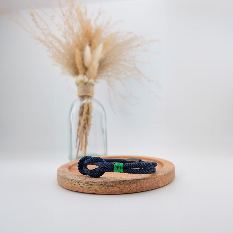 Bracelet cordon bleu marine avec nœud marin et détail tricolore vert, blanc, rouge, posé sur un socle en bois clair.