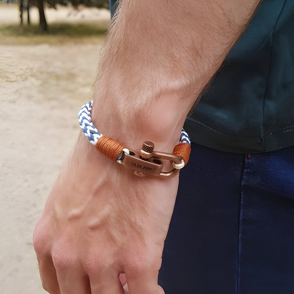 Bracelet cordon bleu et blanc avec fermoir mousqueton ancien en métal cuivré et détails en cuir marron sur poignet masculin.