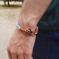 Bracelet cordon bleu et blanc avec fermoir mousqueton ancien en métal cuivré et détails en cuir marron sur poignet masculin.
