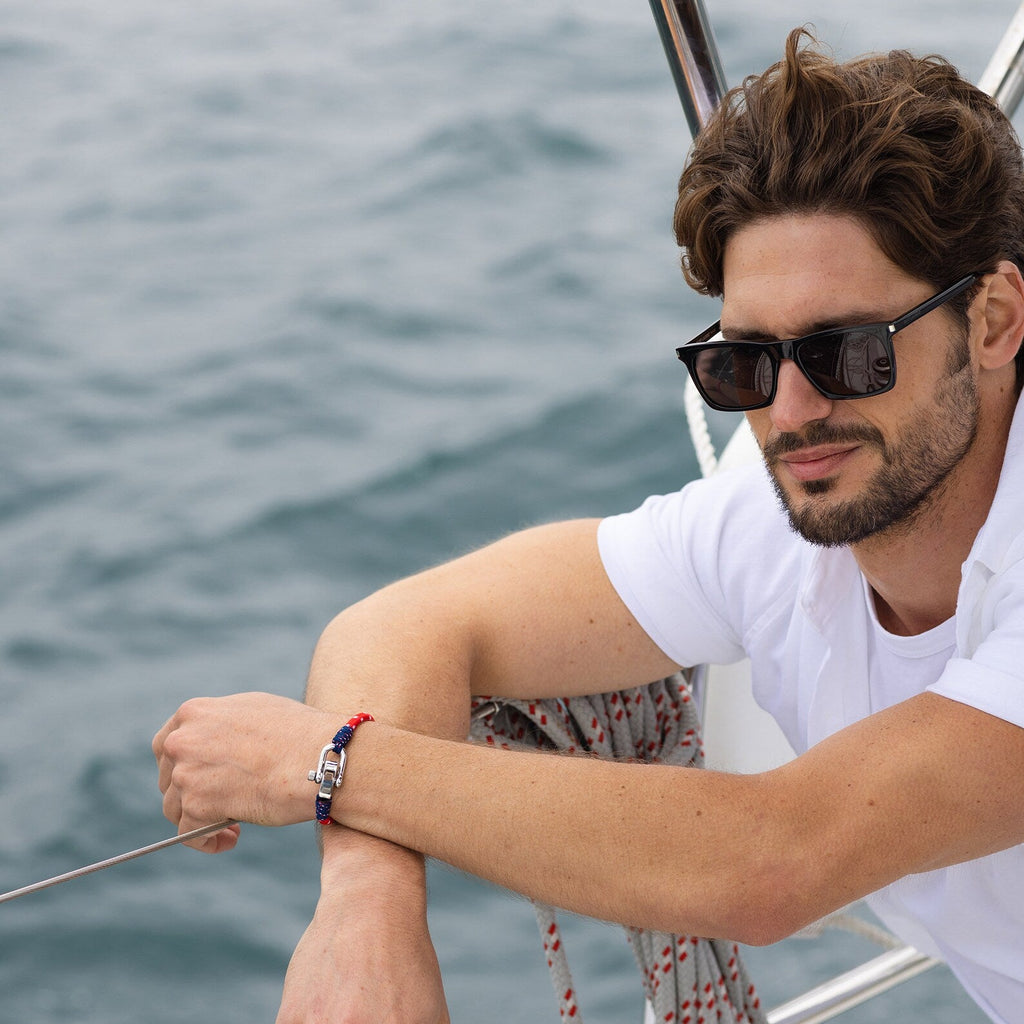 Bracelet cordon bleu, rouge et blanc avec fermoir mousqueton en métal sur poignet d'homme à lunettes noires.