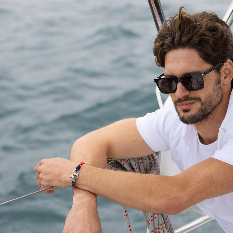 Bracelet cordon bleu, rouge et blanc avec fermoir mousqueton en métal sur poignet d'homme à lunettes noires.