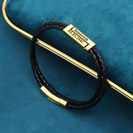 Bracelet cordon noir en cuir tressé avec plaque argentée gravée et fermoir magnétique doré.
