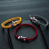 Trois bracelets cordon homme avec manilles acier inoxydable, couleurs jaune, bordeaux et noir, sur surface texturée noire.