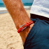 Bracelet cordon orange vif avec mousqueton argenté, style marin, porté au poignet homme sur plage.
