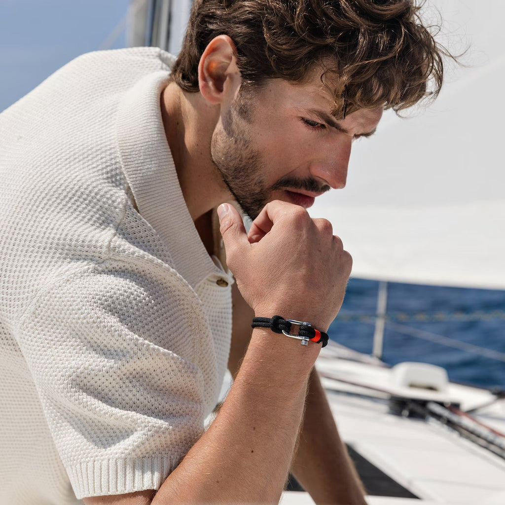 Bracelet cordon noir avec mousqueton argenté porté par un homme au bord d'un voilier.