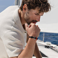 Bracelet cordon noir avec mousqueton argenté porté par un homme au bord d'un voilier.